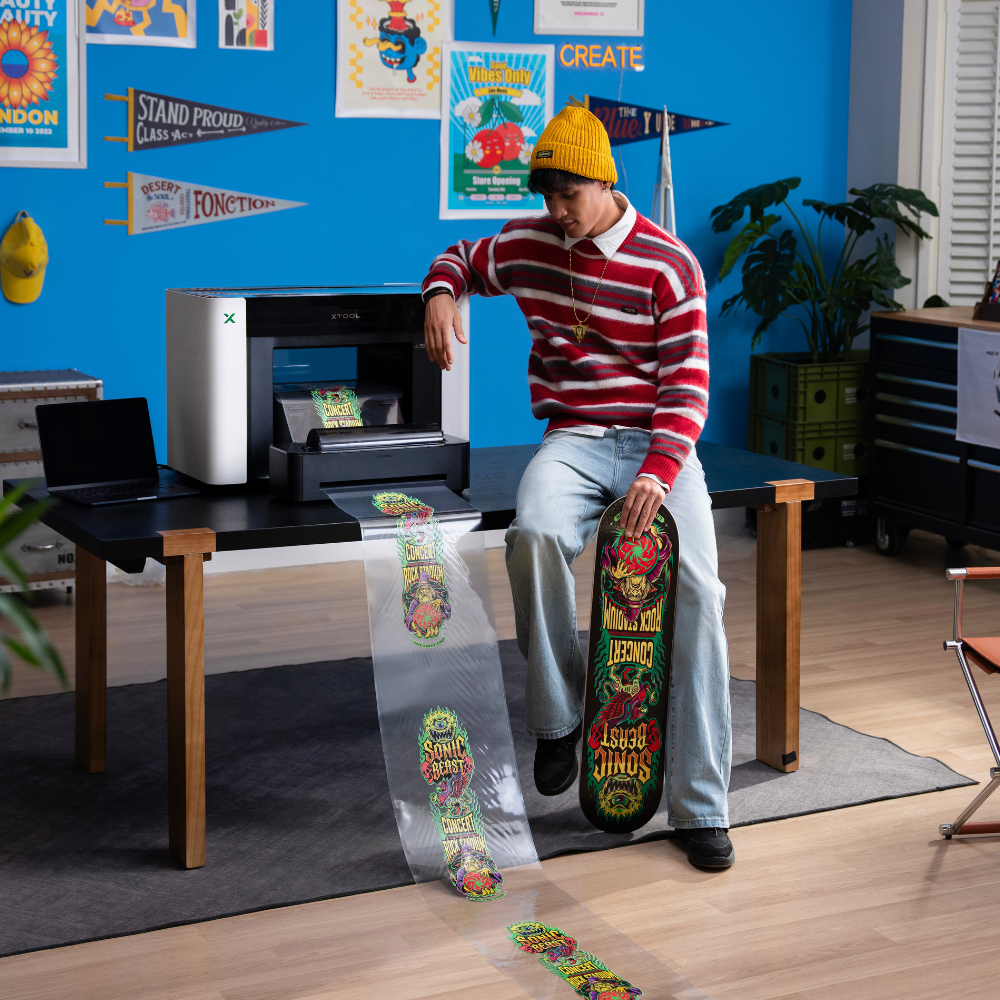 Ein Mann sitzt neben dem xTool O1 Omni Printer und schaut auf einen mehreren Meter langen Folienausdruck für sein Skateboard