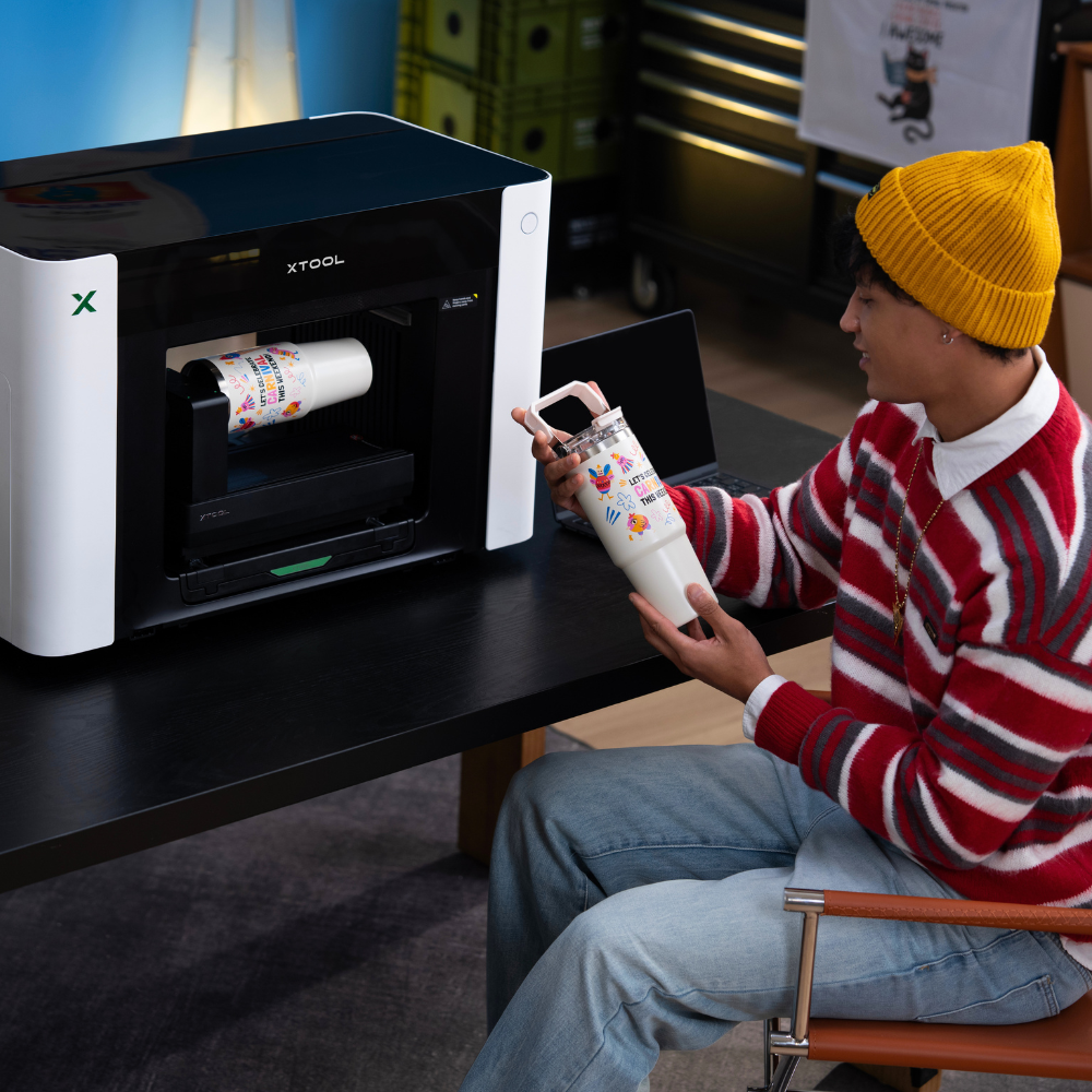Ein Mann sitzt vor dem xTool O1 Omni Printer und hält eine Trinkflasche in der Hand, die er gerade mit dem Gerät bedruckt hat.
