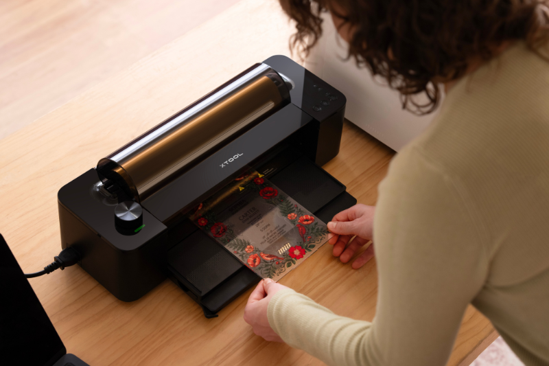xtool-omni-zubehoer-laminator Eine Frau steht vor dem xTool Laminiergerät für den O1 Omni Printer und legt eine bedruckte Acrylplatte ein.