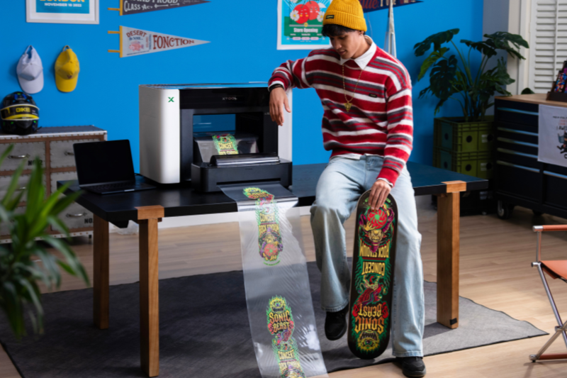 xtool-omni-zubehoer-rollenzufuehrung Ein Mann sitzt neben dem xTool O1 Omni Printer und schaut auf einen mehreren Meter langen Folienausdruck für sein Skateboard