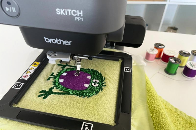 Das Bild zeigt ein Handtuch, auf das ein grünes Monster mit einer Stickmaschine gestickt wird.
